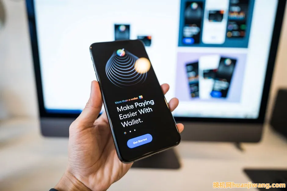 Hand holding smartphone displaying digital wallet app interface, blurred monitor in background.
